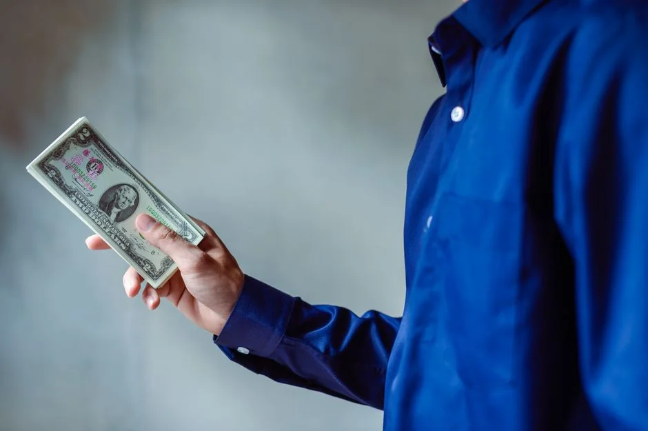 Person in blue shirt holds a stack of US dollar bills, representing GTM engineering salary earnings in 2025.