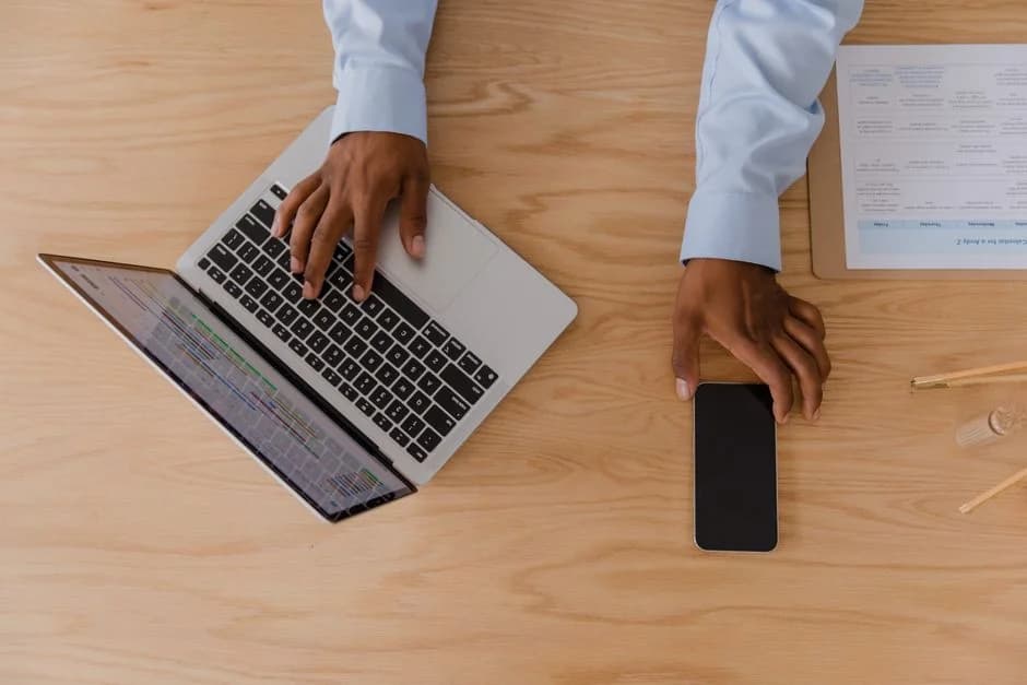 Top-down view of hands working on a laptop and smartphone at a wooden desk, showing multitasking in a modern workspace.