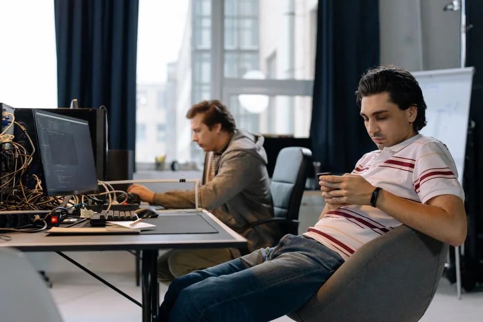 Two male engineers working side by side in a modern office, one at a laptop and the other on a smartphone.